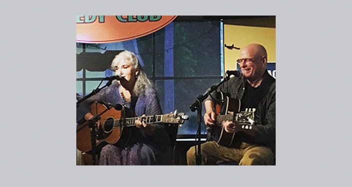 image of local musicians, Lis Williamson and Jim Quine, sitting, playing guitars and singing
