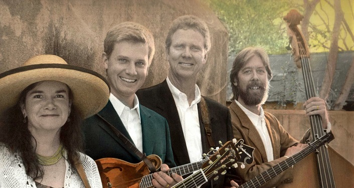 image of The Driftwoods; woman with long dark hair wearing wide-brimmed hat, white dress and holding a guitar; young man to her left wearing white shirt, green jacket holding guitar; to his left middle-aged man wearing white shirt, dark brown jacket holding banjo; to his left another middle-aged man with mustache and beard, wearing white shirt, light brown jacket and holding skinny bass
