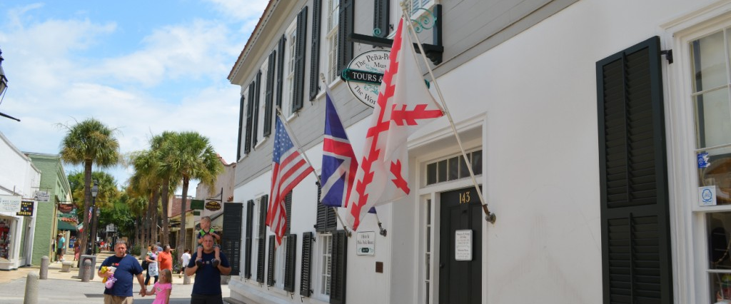 Image contains the side of a house with three flags hanging from it and two adults and one child walking by. Image is of the outdoors.