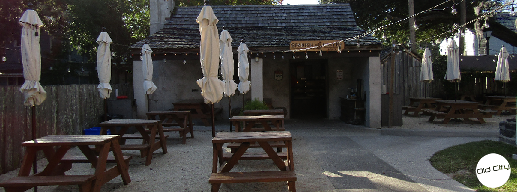 Image contains an outdoor patio with picnic tables, umbrellas, and strings of light in front of a small wooden shop.