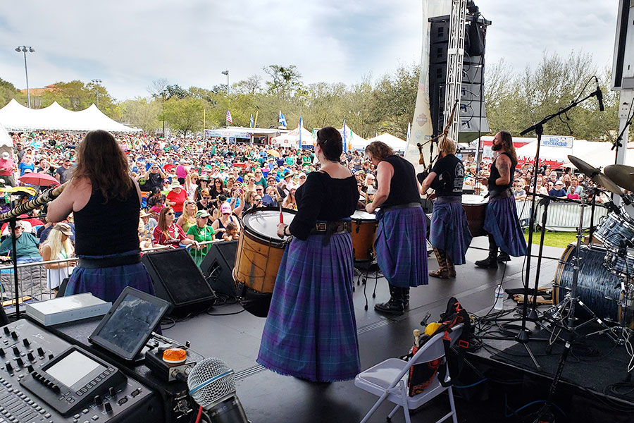 Image is outdoors, and contains a band of five people playing the drums and singing in front of a crowd of hundreds of people.