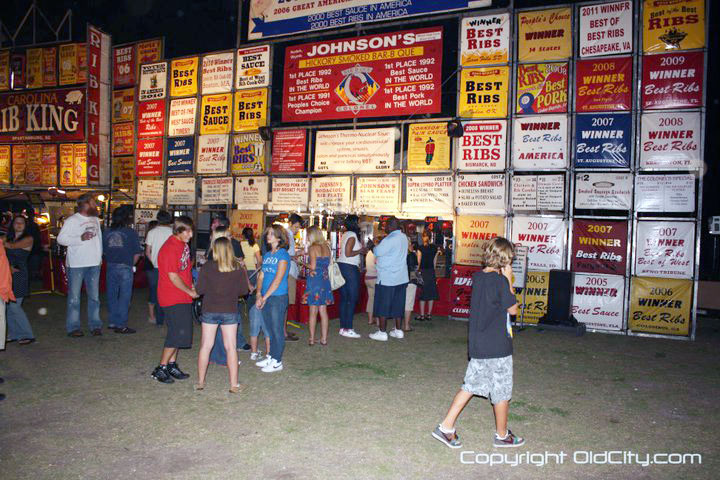 A group of people standing outside, in front of a tall fence with signs on it at Rhythm & Ribs Festival