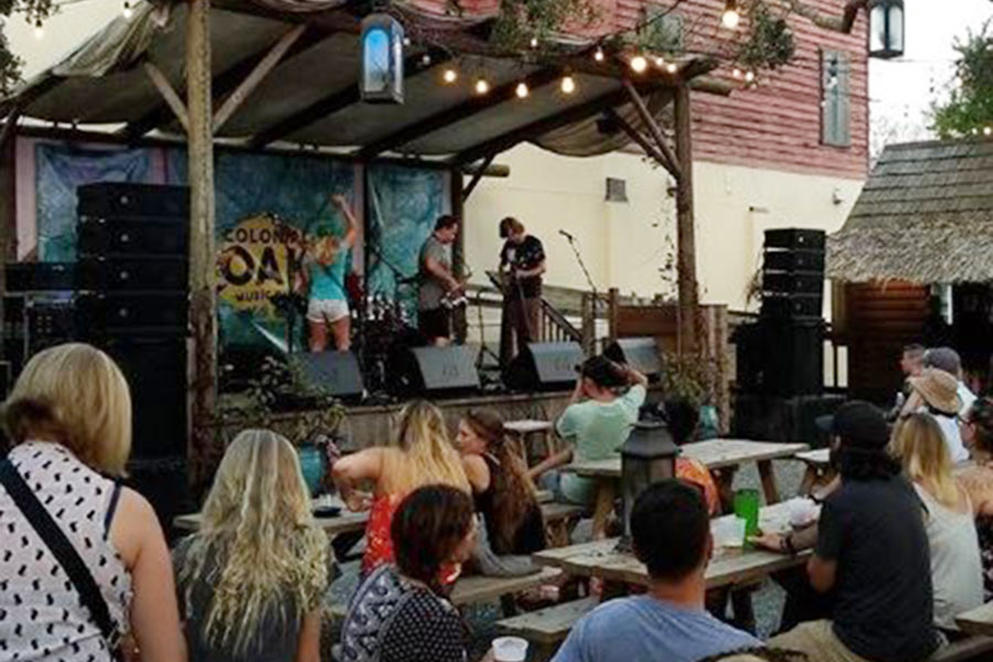 Three people on a stage playing instruments and singing while a crowd of people sit, stand around and watch.