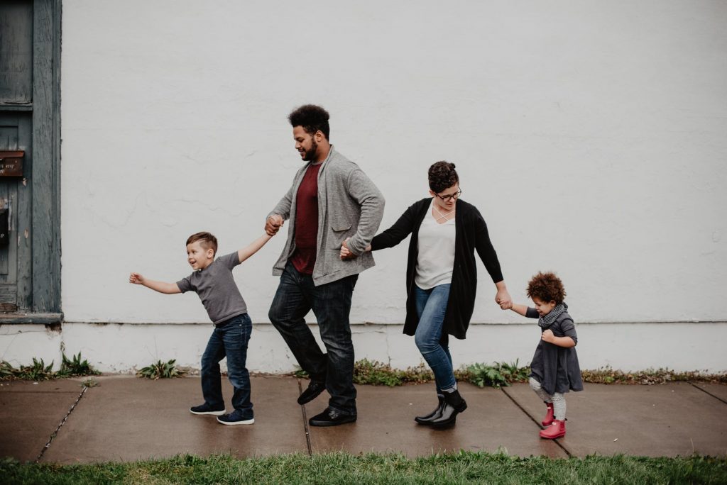 Image contains a mom, dad, and two children holding hands on a sidewalk.