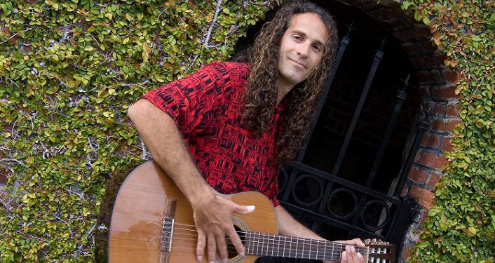 press photo of Fermin Spanish Guitar; long-haired, middle-aged man wearing short-sleeved red print shirt, standing strumming a guitar