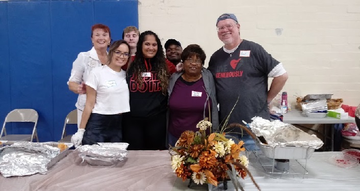 photo of 7 people; four women, 1 younger man, 1 middle-aged - all standing behind table with trays of food and Thanksgving flower arrangement