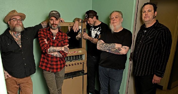 press photo of Americana group Lucero; 5 men all standing in casual poses, 2 with arms crossed. 2 wearing baseball caps, 4 wearing black shirts, 1 wearing red plaid shirt