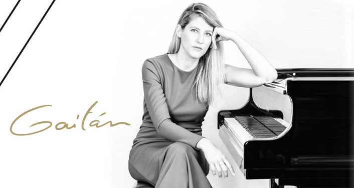 black & white image of woman with long hair, wearing a gown, sitting on piano bench with left arm propped on piano; Internationally acclaimed pianist, Maria Dolores Gaitán