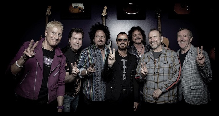 press photo of Ringo Starr and His All Starr Band; six middle-aged men standing looking into camera. Former Beatle, Ringo is making peace sign with right hand.