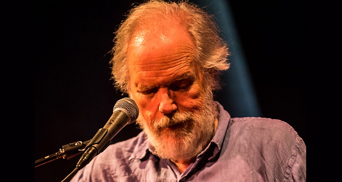 image of Leo Kottke, an older man with beard, slightly balding, looking down, playing a guitar