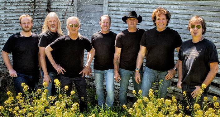 press photo of Southern country-rock legends, The Outlaws; 7 older men standing outside in front of a buildiing in the daytime; all wearing black t-shirts, some black slacks, some blue jeans