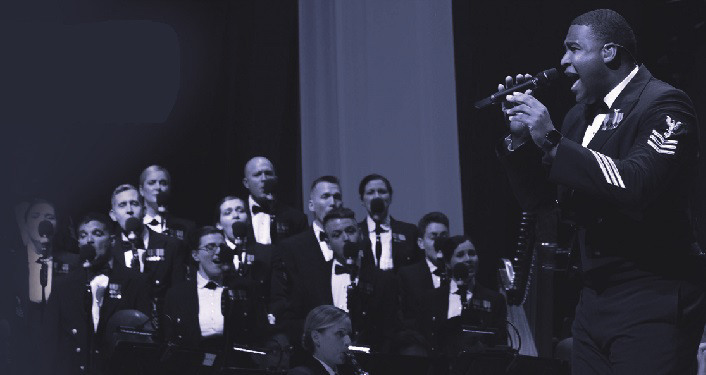 image of US Navy Band in Concert; one man in uniform standing signing into microphone, several others sitting in orchestra in uniform with their insturments