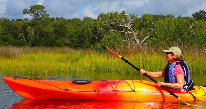side-view of woman in an orange kayak wearing pink shirt, blue vest, and ball cap, kayaking on Moultrie Creek